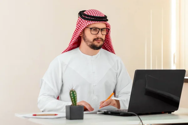 An Arab man, a businessman, a sheikh in a national Arab costume works at a table in the office. Investments, business, work via the Internet, online contracts