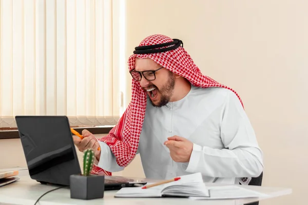 An Arab man, a businessman, a sheikh in a national Arab costume works at a table in the office. Investments, business, work via the Internet, online contracts