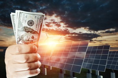 Güneş panellerinin arka planında dolar olan bir el. Güneş paneli maliyeti, güneş enerjili iş. Yeşil elektriğin maliyeti, geleceğin enerjisi.