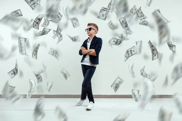 A cute, rich boy with glasses holds dollars and knows how to make money against the background of falling banknotes. Money rain, Financial education, young millionaire, investments, loans, winnings
