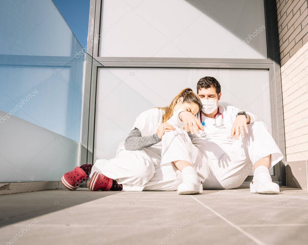 Ground level of tired male and female medics in uniform and masks sitting on floor on balcony and sleeping after hard working day during COVID 19 epidemic