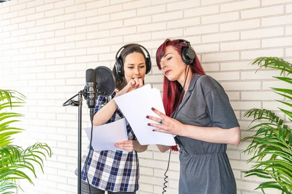 Young women in headphones discussing script while standing near microphone against brick wall in film dubbing studio