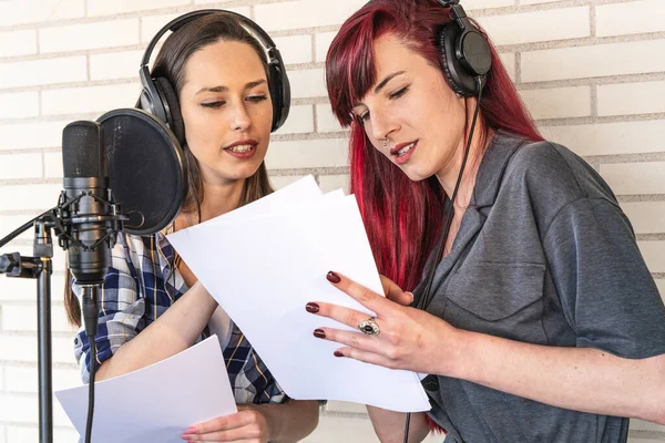 Young women in headphones talking about script while standing near microphone before radio program broadcast in studio