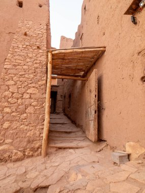 Geleneksel Berber kaşbanın yüksek toprak duvarları arasındaki tipik dar geçitte eski ahşap bir kapı, kırsal sazlık tente ve taştan döşenmiş zemin yer alıyor. Otantik güney Fas mimarisi.