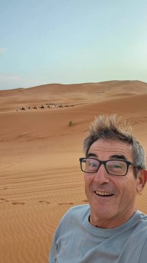 Gözlüklü mutlu yaşlı adam ön planda selfie çekerken uzak bir turist kuyruğu Fas Sahra Sahra 'sında alacakaranlıkta altın kum tepelerini geçiyor..