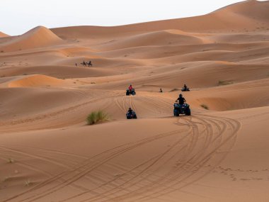 Küçük bir deve kervanı Fas Sahra Sahra 'sında bulutlu bir gökyüzünün altında kum tepelerini geçerken, bir grup turist kumlu yamaçlarda yarışıyor..