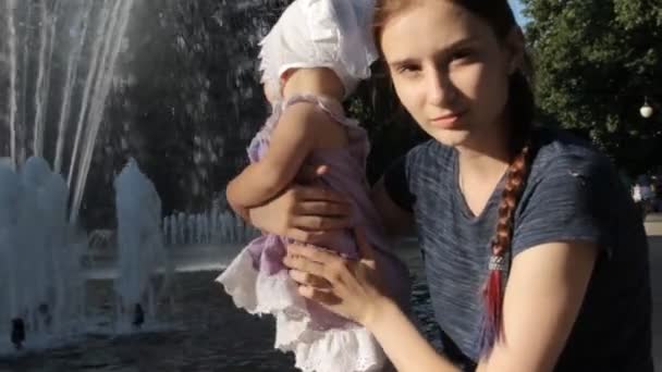 Una niñera joven sosteniendo a un bebé junto a una fuente — Vídeo de stock