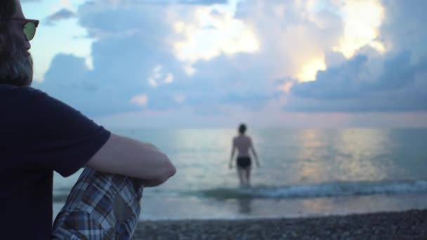 Bearded middle-aged man on beach seashore against background of blured woman in swimsuit setting into sea at sunset — стоковое видео