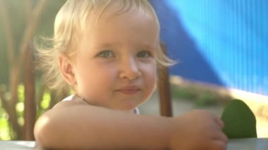 Küçük sarışın bebek elinde büyük yeşil bir yaprak tutuyor güneş ışığında kameraya bakıyor yazın avluda masada oturuyor.