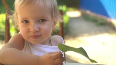 Büyük yeşil yapraklı küçük sarışın bebek güneş ışığında kameraya bakıyor yazın bahçede masada oturuyor.