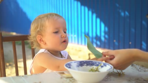 Niña rubia comiendo uvas, recoge gran hoja verde con la mano, jugando y sentado a la mesa en el patio de la casa en verano — Vídeo de stock