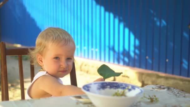 Дитяча дівчинка блондинка їсть виноград, сидячи за столом з великим зеленим листям в руці Літо — стокове відео
