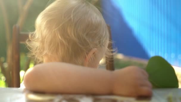 Kleines blondes Baby blickt im Sonnenlicht in die Kamera und sitzt im Sommer am Tisch im Hof — Stockvideo