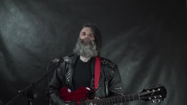 Бородатый рок-музыкант в кожаной куртке с красной электрогитарой, держащей кулак — стоковое видео