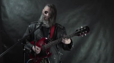 Tıraşlı şakakları, yuvarlak gözlükleri ve deri ceketi olan sakallı kıllı rock 'çı kırmızı elektro gitar çalıyor ve gece kulübü konser sahnesinde siyah perde arkasında şarkı söylüyor.