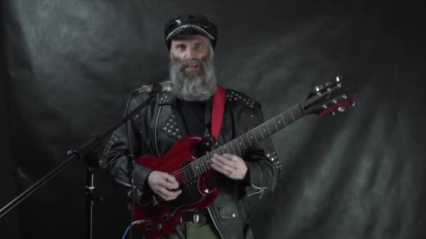 Бородатий рок-музикант у шкіряній куртці та кепці грає на червоній електрогітарі та співає на сцені концерту нічного клубу чорна завіса фону — стокове відео