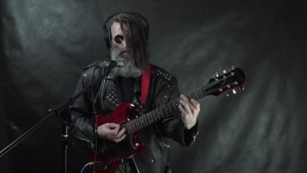 Rocker barbu poilu dans des écouteurs avec temples rasés, lunettes rondes et veste en cuir joue de la guitare électrique rouge, chante et enregistre sa composition en studio — Video