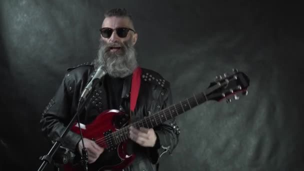 Rocker peludo barbudo en gafas de sol con borde de cuerno retro con templos afeitados y chaqueta de cuero toca la guitarra eléctrica roja en el escenario del club nocturno en concierto con fondo de cortina negro — Vídeos de Stock