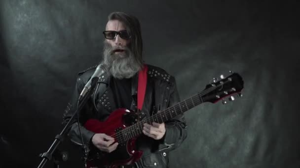 Rocker peludo barbudo en gafas de sol negras retro con sienes afeitadas y chaqueta de cuero toca la guitarra eléctrica roja en el escenario del club nocturno en concierto con fondo de cortina negra — Vídeos de Stock