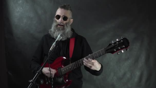 Siyah kapüşonlu ve yuvarlak gözlüklü sakallı rockçı kırmızı elektro gitar çalar, siyah perde arkası ile gece kulübü konserinde şarkı söyler. — Stok video