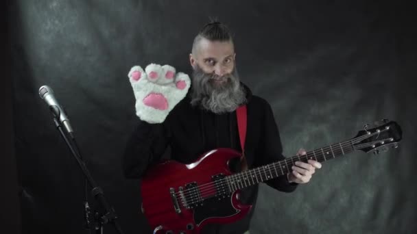 Balancim barbudo com templos raspados em capuz preto sorrindo e acenando para o público com luva de pata de gato branco no concerto de palco da boate com fundo de cortina preta. — Vídeo de Stock