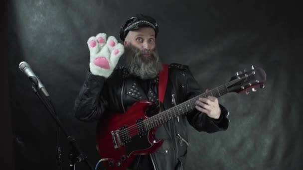 Baard rocker in lederen jas en pet lachend en zwaaiend naar publiek met witte kattenpoot handschoen met rode elektrische gitaar op nachtclub podium concert met zwart gordijn achtergrond. — Stockvideo