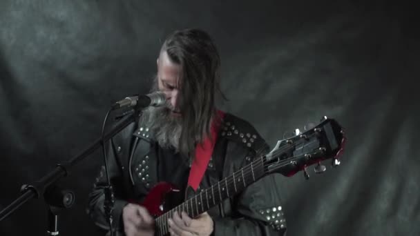 Musicien barbu de rock aux cheveux longs en veste de cuir joue de la guitare électrique rouge et chante sur scène avec un fond noir — Video