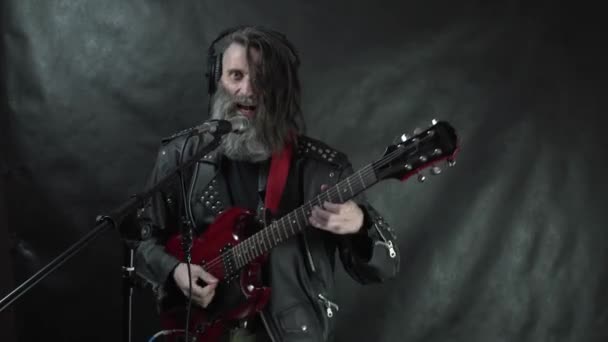 Rockstar barbu d'écouteurs avec temples rasés et veste en cuir joue de la guitare électrique rouge, chante et enregistre sa composition en studio — Video