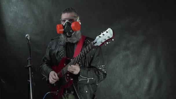 Baard rockster in transparant masker, respirator en lederen jas speelt rode elektrische gitaar op het podium met zwarte achtergrond — Stockvideo