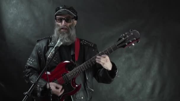 Бородата рок-зірка в ретро чорних сонцезахисних окулярах з голими храмами і шкіряна куртка грає на червоній електрогітарі на сцені з чорним тлом — стокове відео