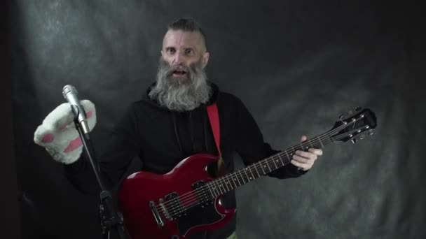 Punk rockster met geschoren tempels, baard en zwarte hoodie speelt door kat poot handschoen op rood elektrische gitaar op het podium met zwarte achtergrond — Stockvideo