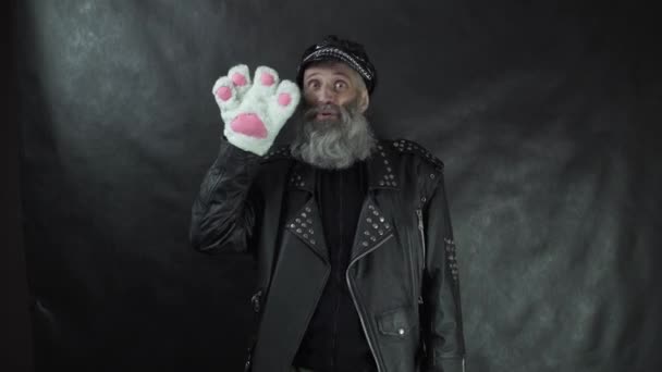 Bärtiger Mann in Lederjacke und Mütze lächelnd und winkend Katzenpfotenhandschuh mit schwarzem Hintergrund — Stockvideo