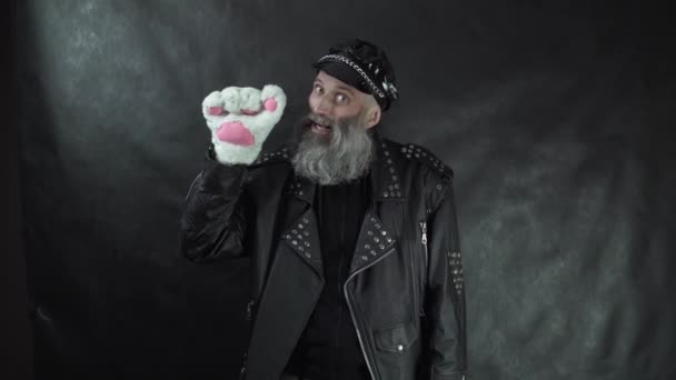 Baard man in lederen jas en pet glimlachend zoet, afscheid nemen en zwaaien kattenpoot handschoen met zwarte achtergrond — Stockvideo