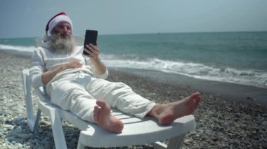 Noel Baba kırmızı şapkalı, beyaz eşofmanlı ve kapüşonlu. Deniz kıyısında akıllı telefon videosu tutuyor. Mutlu Noeller dileyen çocukla konuşuyor.