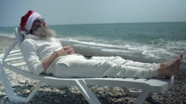 Deniz kıyısında akıllı telefonlu Noel Baba Mutlu Noeller diliyor ve güneş gözlüğü takan yeni yıl güneşleniyor..
