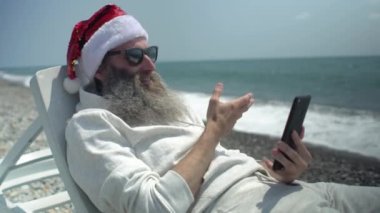 Kırmızı şapka takan ve güneş gözlüğü takan Noel Baba deniz kıyısını anlatıyor ve Mutlu Noeller diliyor. İnternetteki akıllı telefondaki video sohbetinde deniz kenarında güneşlenirken mutlu yıllar diliyor..