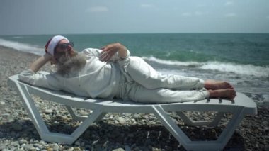 Deniz kıyısında kırmızı şapkalı ve güneş gözlüklü Noel Baba güneşleniyor, elini sallayarak serinliyor, herkesi selamlıyor ve kameraya baş parmağını gösteriyor.