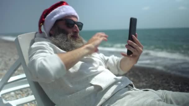 Großaufnahme Weihnachtsmann mit rotem Hut und Sonnenbrille singt Weihnachtslied in virtuellem Video-Online-Chat auf Smartphone sitzend auf Sonnenliege am Meer. — Stockvideo