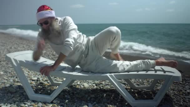 Der Weihnachtsmann am Meer mit rotem Hut und Sonnenbrille bräunt sich sonnengebräunt und dreht sich auf der anderen Seite auf einer Liege liegend um — Stockvideo