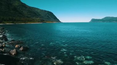 Kayalık bir sahil üzerinde yavaş ve alçak bir uçuş açık, masmavi bir su ve güneşli bir hava. Arka planda deniz ve dağlar var. Norveç 2020.