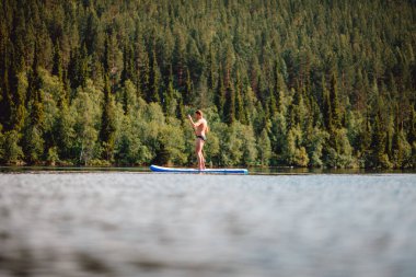 Kürek çeken bir adam ormanın arka planında, mavi göl suyuyla kürek çekiyor.