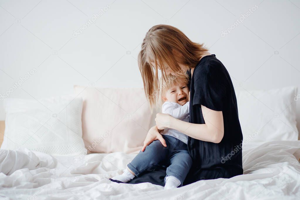 Baby girl experiences colic and pain, young woman mom calms and comforts child hugs, light background bedroom