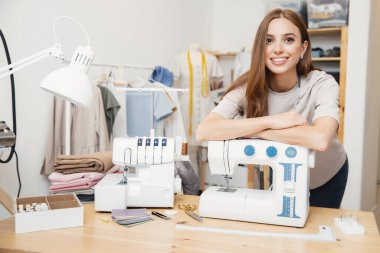 Küçük işletme ev kıyafetleri sipariş konsepti. Kadın terzi kıyafetleri gülümsüyor, dikiş makinesine yaslanıyor.