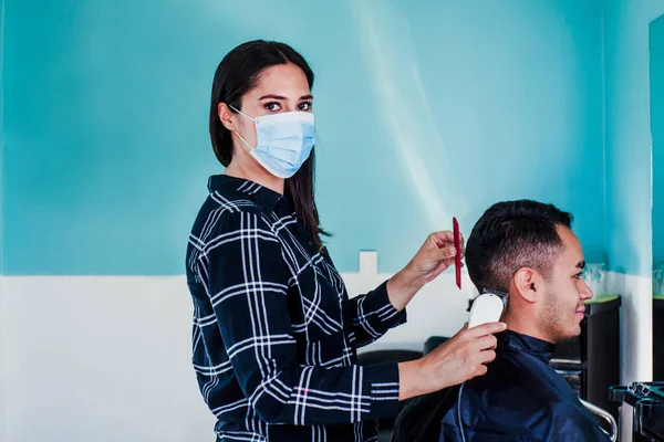 latin woman hairdresser with face mask cutting hair with clipper machine and comb. Mexican man gets a haircut. Barber services with security measures from Covid-19 in Mexico city