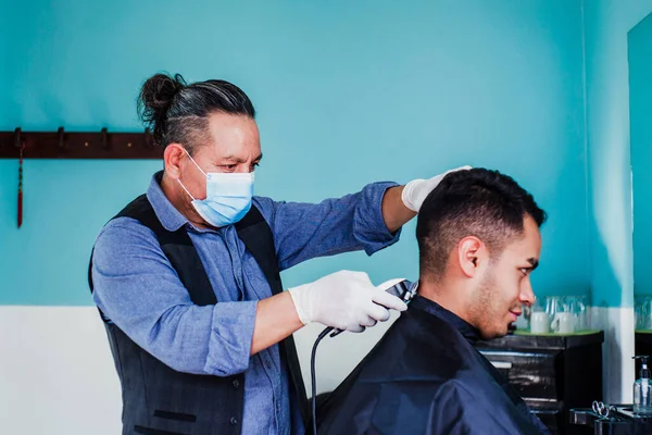 latin man stylist with facemask cutting hair to a client in a barber shop in Mexico city in coronavirus pandemic