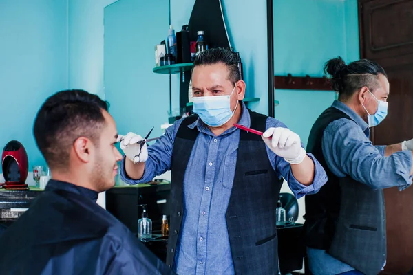 latin man stylist with facemask cutting hair to a client in a barber shop in Mexico city in coronavirus pandemic