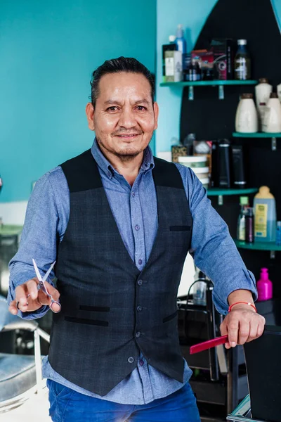 Portrait of latin man barber holding equipments in hand, looking at camera in a barber shop small business in Mexico city