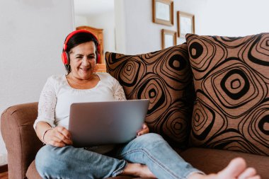Latin, olgun bir kadın, elinde akıllı telefonuyla kablosuz kulaklık takıyor ve Mexico City 'deki evinde internet müziği dinliyor.
