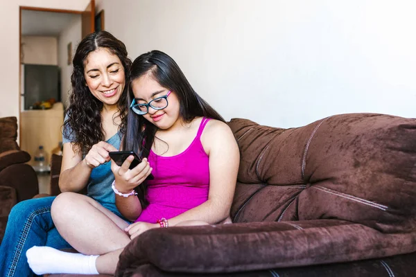 Latin woman with her teenage daughter with down syndrome sitting on the sofa using the phone, in disability concept in Latin America