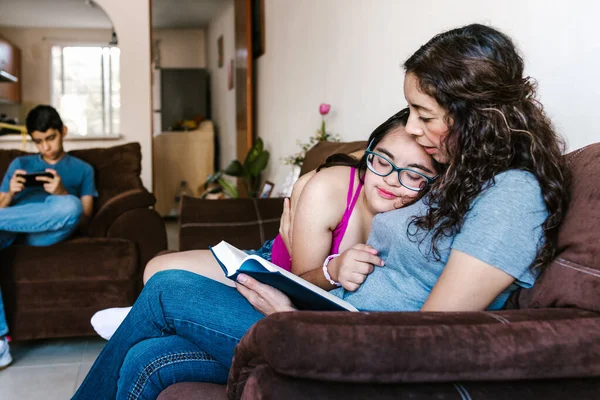 Latin mother reading a book to her teenage daughter with down syndrome sitting on the sofa at home, in disability concept in Latin America